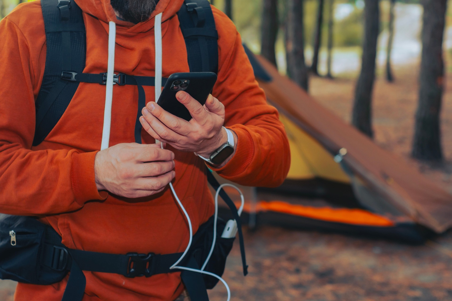 Close-up of a man with a hiking backpack and a smartphone and portable power bank in hands, against the background of a tent in the forest. Digital nomad's concept of sustainable existence in nature.